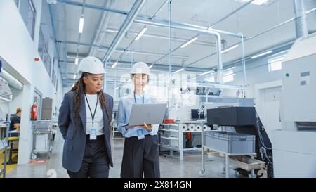 Two Female Heavy Industry Employees Wearing Hard Hats at Factory. Walking and Discussing Industrial Machine Facility, Using Laptop Computer. Asian Engineer and African American Technician at Work. Stock Photo