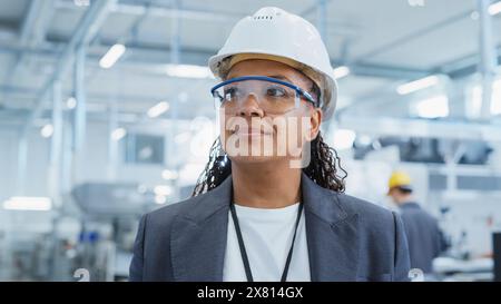 Close Up Portrait of a Happy and Smiling Middle Aged African Female Engineer in White Hard Hat Standing at Electronics Manufacturing Factory. Black Heavy Industry Specialist Supervising Work. Stock Photo