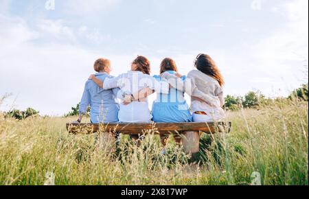 Four smiling women dressed light summer clothes harm embracing sitting on the meadow bench during outdoor walking. Woman's friendship, relations, and Stock Photo