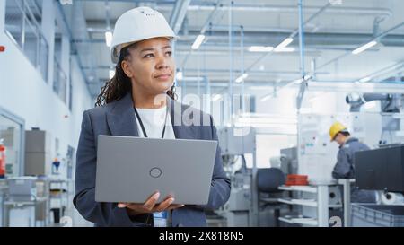 Black African Female Engineer in Hard Hat Standing and Using Laptop Computer at Electronic Manufacturing Factory. Technician Working on Daily Tasks and Research and Development Data. Stock Photo