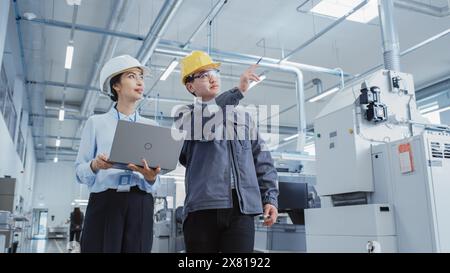 Two Heavy Industry Employees in Hard Hats Discussing Job Assignments at the Factory, Using Laptop Computer. Asian Engineer and Technician at Work Checking Safety Procedures. Stock Photo