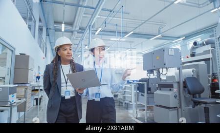 Two Professional Heavy Industry Employees in Hard Hats at Factory. Checking and Discussing Industrial Facility, Working on Laptop Computer. African American Engineer and Asian Technician at Work. Stock Photo