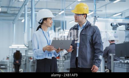 Two Diverse Young Heavy Industry Engineers in Hard Hats Walking with Laptop Computer and Talking in a Factory. Two Manufacturing Employees at Work in Research and Development Facility. Stock Photo