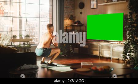 Home Training: Handsome Muscular Black Man Doing Squats, Exercising with Trainer via TV Online Video App. Strong Mixed Race Sportsman Using Chroma Key Green Screen for Virtual Gym. Stock Photo