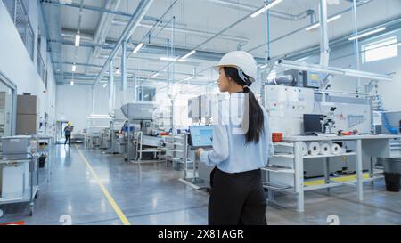 Research and Development Specialist in a Factory Facility, Holding Laptop Computer and Walking in Heavy Industry Machine. Asian Quality Control Manager Investigating the Work Space. Stock Photo