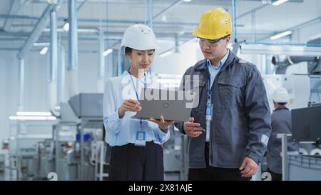 Portrait of Two Young Asian Heavy Industry Engineers in Hard Hats Standing with Laptop Computer and Discussing Work Process in a Factory. Two Manufacturing Employees Chatting in Production Facility. Stock Photo