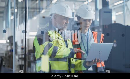 Two Professional Heavy Industry Engineers Wearing Safety Uniform and Hard Hats Discussing Industrial Machine Part on Laptop Computer. Asian Specialist and Middle Aged Technician at Work. Stock Photo