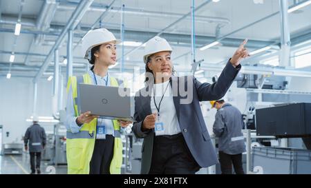 Portrait of Two Female Employees in Hard Hats at Factory. Discussing Job Assignments at Industrial Machine Facility, Using Laptop Computer. Asian Engineer and African American Technician at Work. Stock Photo