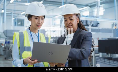 Portrait of Two Female Employees in Hard Hats at Factory Discussing Assignments at Industrial Machine Facility, Using Laptop Computer. Smiling Asian Engineer and African American Technician at Work. Stock Photo