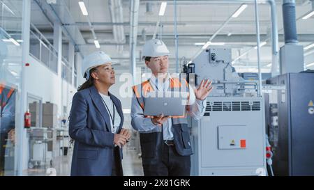 Two Professional Heavy Industry Employees in Hard Hats at Factory. Checking and Discussing Industrial Facility, Working on Laptop Computer. African American Engineer and Asian Technician at Work. Stock Photo