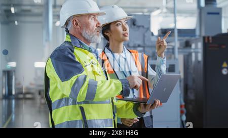 Two Diverse Professional Heavy Industry Engineers Wearing Safety Uniform and Hard Hats, Working on Laptop Computer. Asian Technician and Worker Talking Next to a Factory Machine. Stock Photo