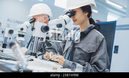 Young Asian Industrial Scientist and Older Engineer Working at a Desk in a Factory Facility, Using Microscopes to Inspect the Manufacturing Production Parts for Quality. Stock Photo