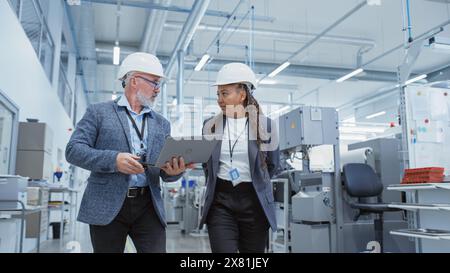 Portrait of a Two Diverse Heavy Industry Engineers in Hard Hats Walking with Laptop Computer and Talking in a Factory. Footage of Two Manufacturing Employees at Work. Stock Photo