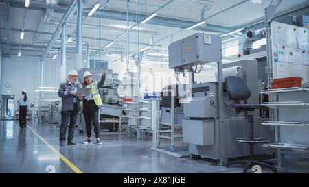 Two Professional Heavy Industry Colleagues in Hard Hats at Factory. Checking and Discussing Industrial Facility, Using Laptop Computer. Middle Aged Engineer and African American Technician at Work. Stock Photo