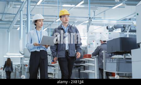Two Young Asian Heavy Industry Engineers in Hard Hats Walking with Laptop Computer and Discussing Work Process in a Factory. Two Manufacturing Employees at Work in Production Facility. Stock Photo