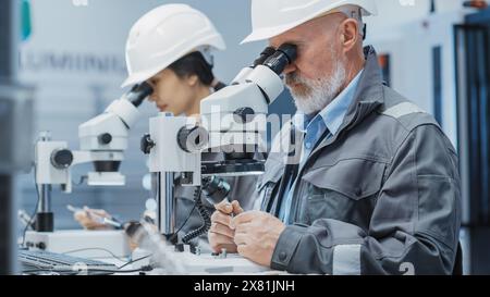 Middle Aged Industrial Scientist and Younger Asian Engineer Working at a Desk in a Factory Facility, Using Microscopes to Work on Small Manufacturing Production Parts and Details. Stock Photo