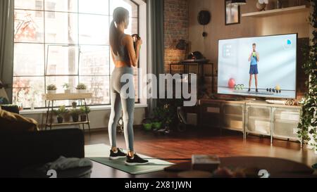 Home Gym: Asian Girl Training Using Kettlebell, Exercising with Personal Trainer via TV Online Video App. Authentic Woman Using Workout Service Fitness Streaming App for Virtual Training Stock Photo