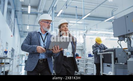 Two Professional Heavy Industry Engineers Wearing Hard Hats at Factory. Walking and Discussing Industrial Machine Facility, Working on Laptop. African American Manager and Technician at Work. Stock Photo