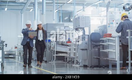 Two Diverse Heavy Industry Engineers in Hard Hats Walking with Laptop Computer and Talking in a Factory. Footage of Two Manufacturing Employees at Work. Stock Photo