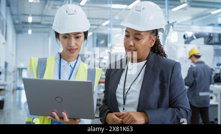 Portrait of Two Female Employees in Hard Hats at Factory Discussing Assignments at Industrial Machine Facility, Using Laptop Computer. Smiling Asian Engineer and African American Technician at Work. Stock Photo