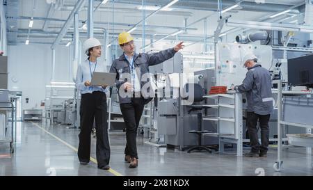Wide Shot: Two Heavy Industry Employees in Hard Hats Discussing Job Assignments at the Factory, Using Laptop Computer. Asian Engineer and Technician at Work Smiling Stock Photo