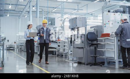 Wide Shot: Two Heavy Industry Employees in Hard Hats Discussing Job Assignments at the Factory, Using Laptop Computer. Asian Engineer and Technician at Work Smiling Stock Photo