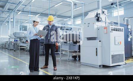 Two Professional Heavy Industry Employees Wearing Hard Hats at Factory. Walking and Discussing Industrial Machine Facility, Using Laptop Computer. Asian Engineer and Technician at Work. Stock Photo