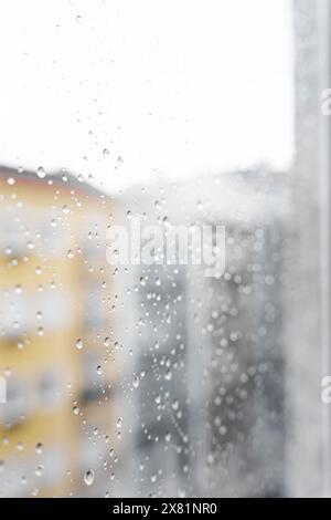 Raindrops on the window glass from the side of the apartment during the day, in the background there are colorful buildings and the street. Stock Photo