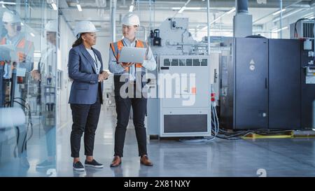 Two Professional Heavy Industry Colleagues in Hard Hats at Factory. Checking and Discussing Industrial Facility, Working on Laptop Computer. African American Engineer and Asian Technician at Work. Stock Photo