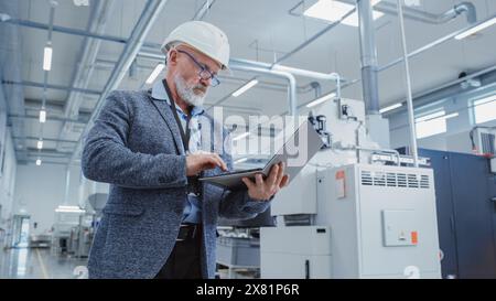Portrait of a Bearded Middle Aged Engineer Standing in a Factory Facility, Wearing Smart Casual Clothes and a White Hard Hat. Heavy Industry Specialist Working on Laptop Computer. Stock Photo