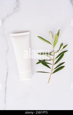 Top view of white plastic tube without label and olive branch on white marble background. Minimal flat lay of bottle of lotion, creme, balsam Stock Photo