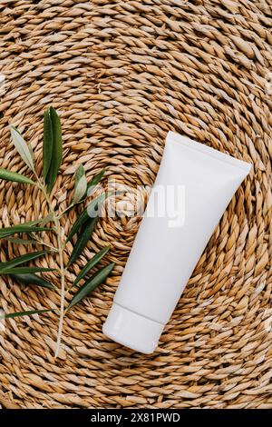 Top view of white plastic tube without label and olive branch on wicker beige background. Minimal flat lay of bottle of lotion, creme, balsam Stock Photo