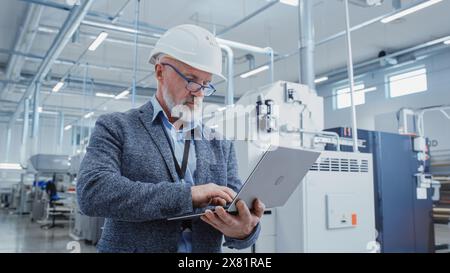 Portrait of a Bearded Middle Aged General Manager Standing in a Factory Facility, Wearing Casual Suit and a White Hard Hat. Heavy Industry Specialist Working on Laptop Computer. Stock Photo
