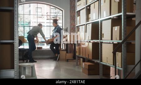 Multicultural Male and Female Warehouse Inventory Managers Talking, Using Laptop Computer and Checking Retail Stock. Rows of Shelves Full of Cardboard Box Packages in the Background. Stock Photo