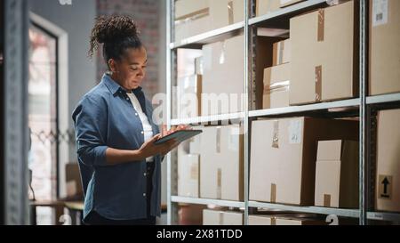 Portrait of a Successful Employee Checking Inventory, Writing in Tablet Computer. Black Woman Posing for Camera and Smiling in a Warehouse Storeroom with Orders Ready for Shipment. Stock Photo