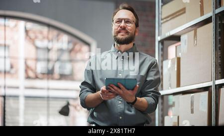Portrait of a Happy Small Warehouse Employee Checking Inventory, Using Tablet. Handsome Man with Moustache Smiling in a Storeroom with Parcels and Online Purchased Orders Ready for Shipment. Stock Photo