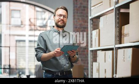 Portrait of a Successful Employee Checking Inventory, Writing in Tablet Computer. Stylish Man Posing for Camera and Smiling in a Warehouse Storeroom with Orders Ready for Shipment. Stock Photo