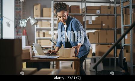 Warehouse Female Worker Using Laptop Computer, Preparing a Small Parcel for Shipping. African American Small Business Owner Working in Storeroom, Preparing Order for Client. Stock Photo