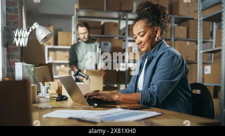 Diverse Male and Female Warehouse Inventory Managers Talking, Using Laptop Computer and Checking Retail Stock. Rows of Shelves Full of Cardboard Box Packages in the Background. Stock Photo