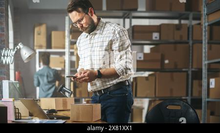 Storeroom Inventory Worker Using Smartphone to Scan a Barcode on Parcel, Preparing a Small Cardboard Box for Postage. Small Business Owner Working on Laptop in Warehouse with Colleague. Stock Photo