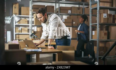 Successful Small Business Owner of a Retail Online Shop Preparing a Small Cardboard Parcel for Postage. Stylish Young Inventory Manager Working on Laptop Computer in Warehouse Facility. Stock Photo