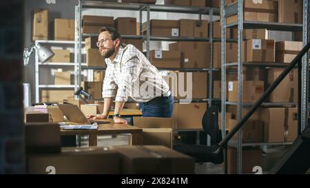 Stylish Small Business Owner Preparing a Small Parcel for Postage. Inventory Manager Thinking About Successful Career, Looking at the Sun from a Window. Working on Laptop in Warehouse Facility. Stock Photo