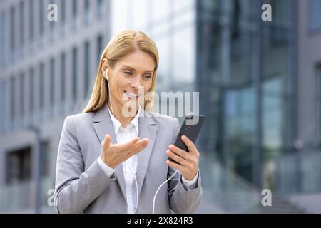 A confident businesswoman in a grey suit talks on her smartphone with earphones outside a modern office building. She showcases a professional and focused demeanor, embodying success in her career Stock Photo