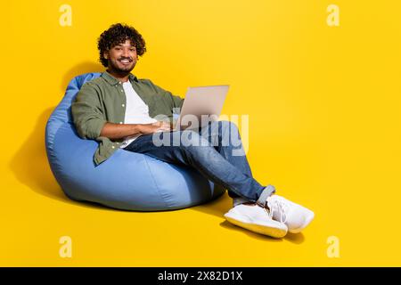Photo of young arab guy remote studying in khaki shirt sitting pouf junior it specialist at lounge isolated on yellow color background Stock Photo