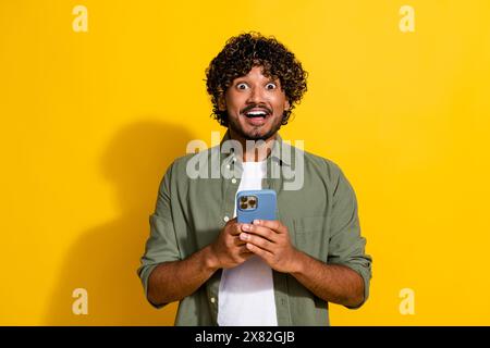 Photo portrait of young funny wavy haired mexican guy using his smartphone shocked browsing news feed isolated on yellow color background Stock Photo