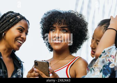 Curly-haired latina woman laughs while using cell phone with her friends. Stock Photo