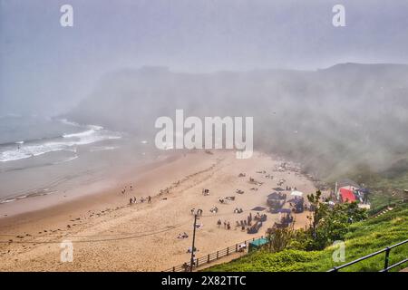 A foggy beach scene with people relaxing on the sand and a few tents set up. The ocean waves are gently crashing onto the shore, and the background is Stock Photo