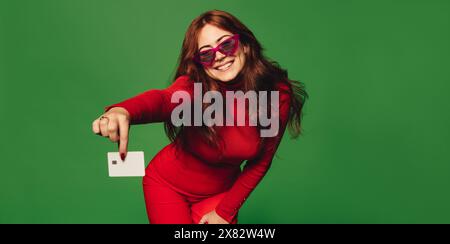 Stylish woman pays with a vibrant red credit card against a green background. She smiles, wearing trendy sunglasses. Stock Photo