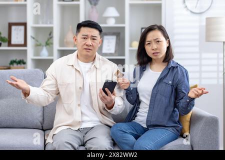 Confused couple sitting on a couch with a smartphone and credit card, facing online payment issues. They appear frustrated and puzzled. Stock Photo