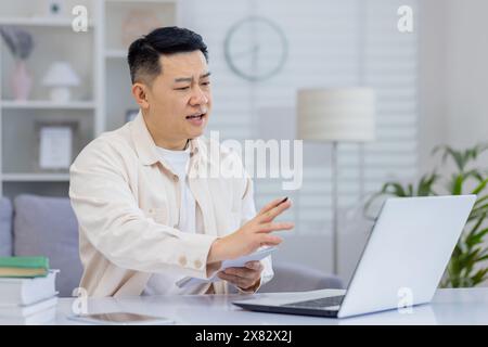 Middle-aged man on a video conference call while working from home office, using a laptop, tablet, and books. Stock Photo